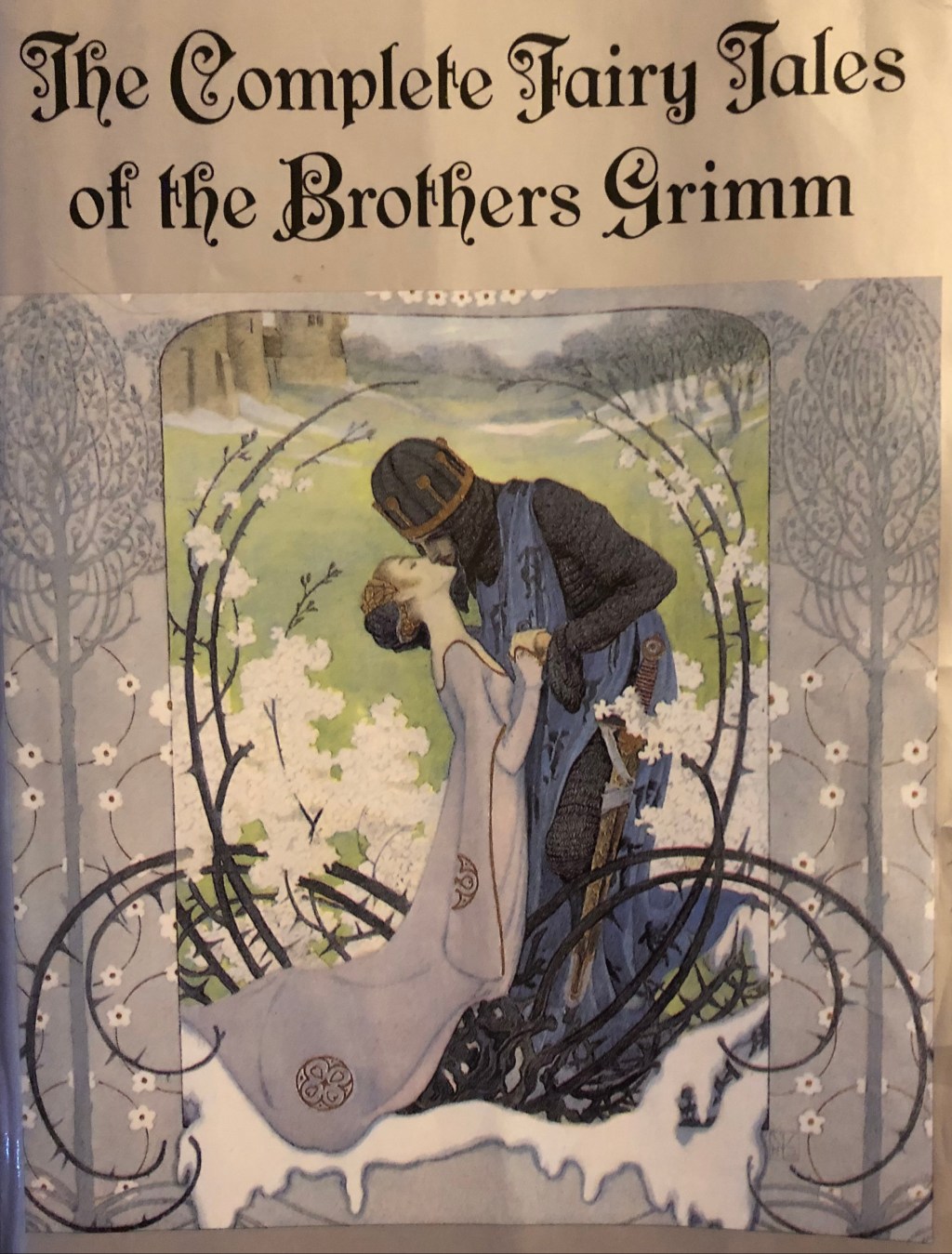 Close up on front cover of somewhat 1992 paperback edition of THE COMPLETE FAIRY TALES OF THE BROTHERS GRIMM, translated and with introduction by Jack Zipes. Book is a pale, silvery purple color and the artwork shows a medieval-esque fairy tale princess in a pale purple gown kissing a prince dressed in chain mail, both encircled by flowering vines.